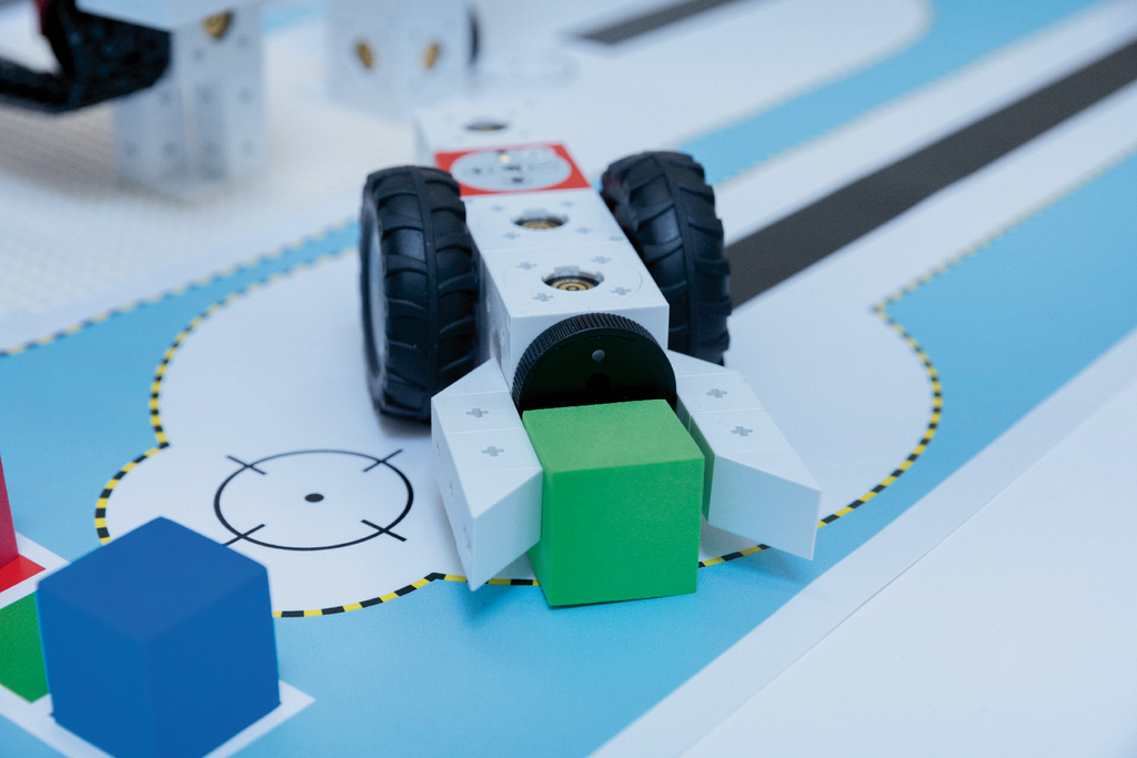 Das Bild zeigt den Lernroboter eXperiBot, der auf einer abgebildeten Lernumgebung platziert ist. Er f&uuml;hrt eine Aufgabe aus, bei der er einen gr&uuml;nen Block aufnimmt und sich dabei zwischen bunten Formen bewegt.