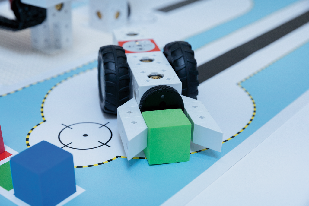 Das Bild zeigt einen Roboter mit gro&szlig;en Reifen, der offenbar einen gr&uuml;nen W&uuml;rfel transportiert. Im Hintergrund ist eine farbige, mit verschiedenen Formen und einer Stra&szlig;e gestaltete Experimentierfl&auml;che zu sehen.