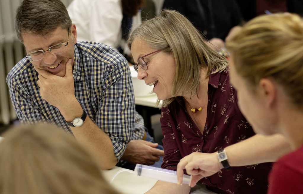 Auf dem Bild sind mehrere Personen zu sehen, die an einem Tisch sitzen und sich aktiv austauschen. Eine Frau zeigt einem Mann ein Experimentiergerät, während beide sichtlich Spaß an der Interaktion haben.