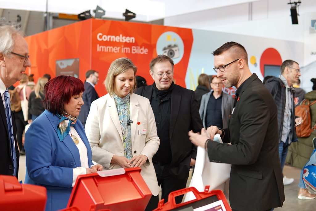 Das Bild zeigt mehrere Personen, die an einem Stand von Cornelsen stehen und Interesse an roten Experimentierkoffern zeigen. Eine Person erklärt anschaulich die Inhalte und Anwendungen der Materialien für das deutsche Bildungswesen.
