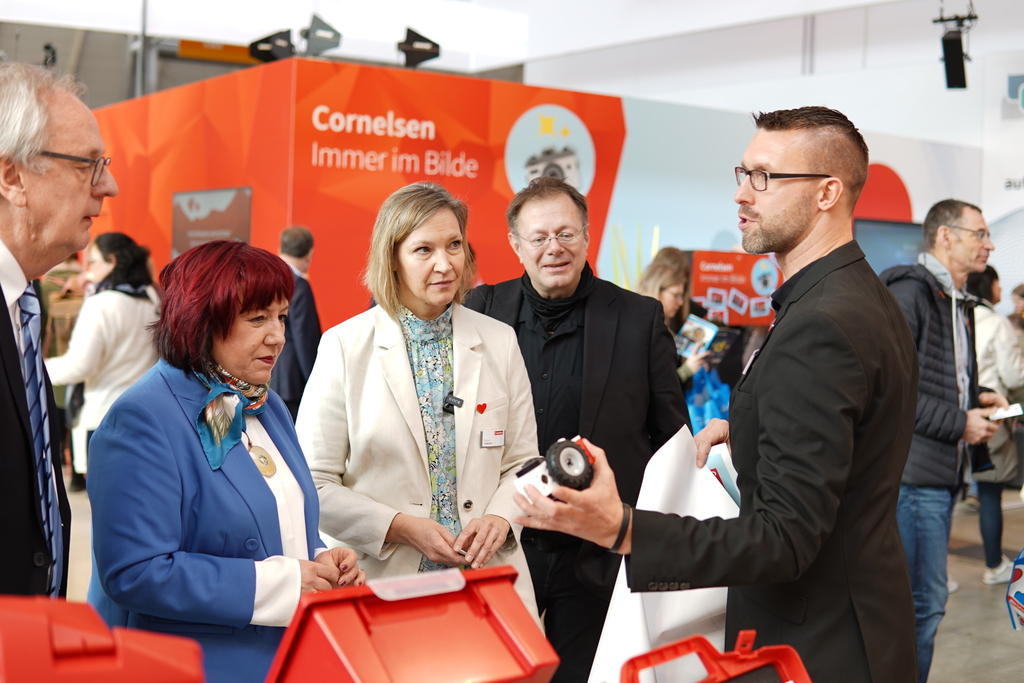 Das Bild zeigt eine Gruppe von Personen, die einem Pr&auml;sentierenden aufmerksam zuh&ouml;ren, w&auml;hrend er ihnen ein Produkt zeigt. Im Hintergrund ist ein auff&auml;lliges, orangefarbenes Banner von Cornelsen zu sehen, das auf deren Engagement im Bildungsbereich hinweist.