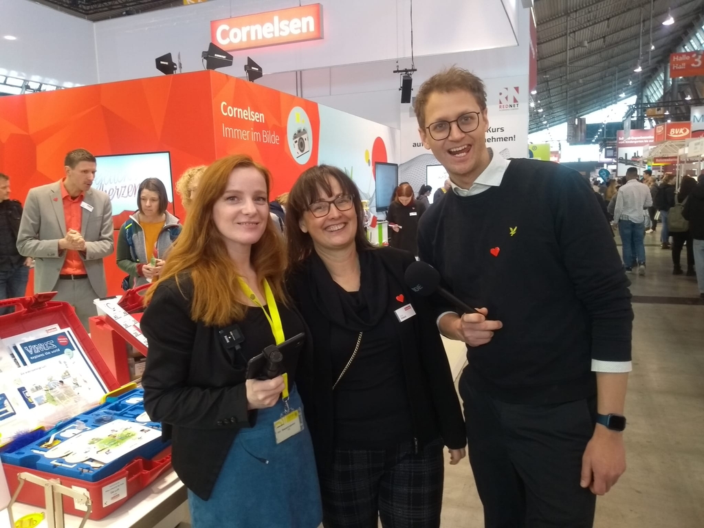 Das Bild zeigt mehrere Personen an einem Messestand von Cornelsen, der sich mit Experimentierbedarf f&uuml;r das Bildungswesen besch&auml;ftigt. Im Vordergrund l&auml;cheln zwei Frauen und ein Mann in formeller Kleidung in die Kamera, w&auml;hrend im Hintergrund weitere Besucher zu sehen sind.