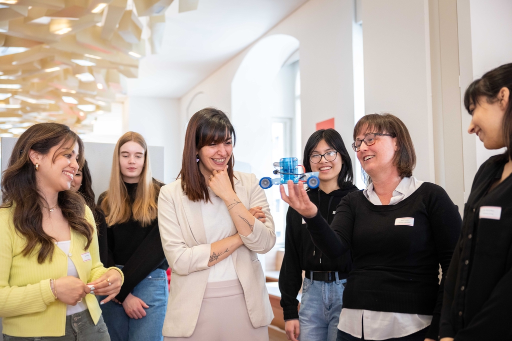 Das Bild zeigt eine Gruppe von Frauen, die aufmerksam auf ein kleines Modell eines Wasserstoffautos schauen, das von einer Frau in der Mitte pr&auml;sentiert wird. Alle wirken interessiert und fr&ouml;hlich, w&auml;hrend sie sich in einem modernen Raum um das Fahrzeug versammelt haben.