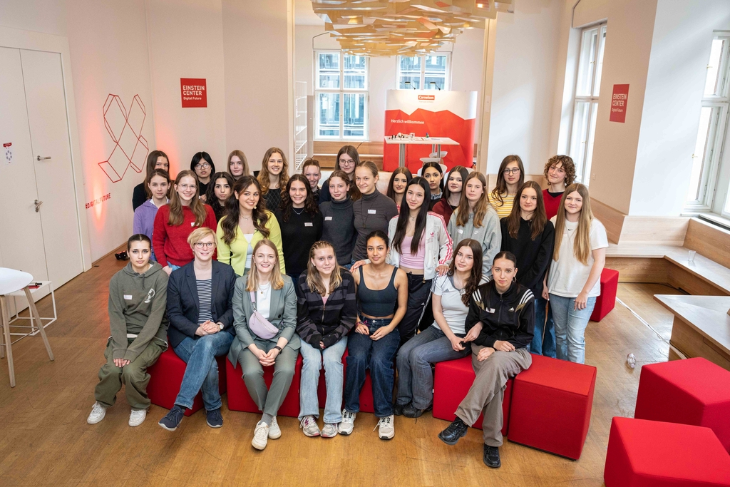 Das Bild zeigt eine Gruppe von jungen Frauen, die in einem modernen Raum mit roten Sitzmöbeln zusammenstehen. Der Hintergrund ist hell gestaltet und enthält das Logo des Einstein Centers, das auf digitale Zukunftstechnologien hinweist.