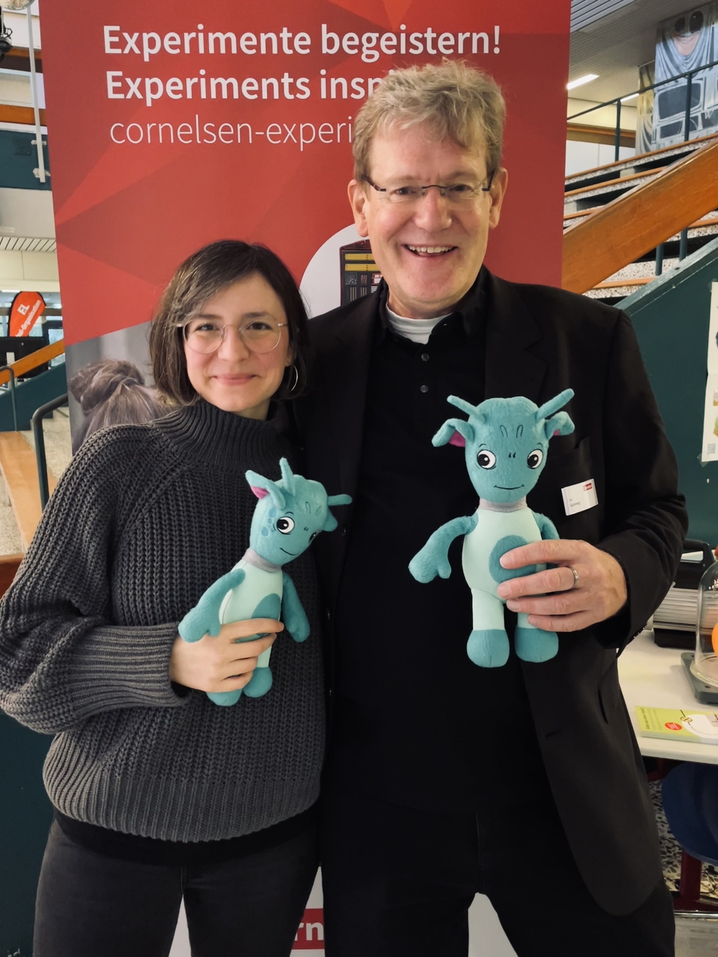 Auf dem Bild stehen zwei Personen vor einem Werbeplakat, das den Slogan "Experimente begeistern!" zeigt. Beide halten den Vinus in der Hand, das eine fantasievolle Kreatur darstellt.