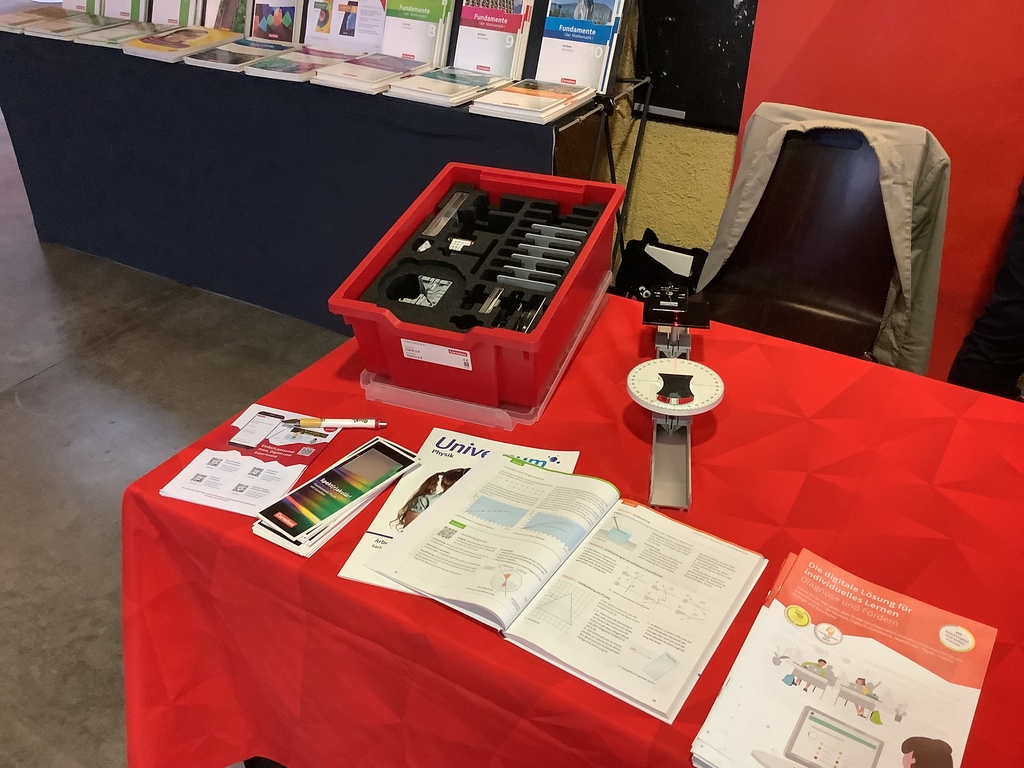 Das Bild zeigt einen Messetisch von Cornelsen Experimenta mit Experimentierbedarf, darunter die rote Experimentier-Wanne des Sch&uuml;ler-Sets Optik 2.0. Zudem sind einige Brosch&uuml;ren und B&uuml;cher &uuml;ber Physik ausgestellt.