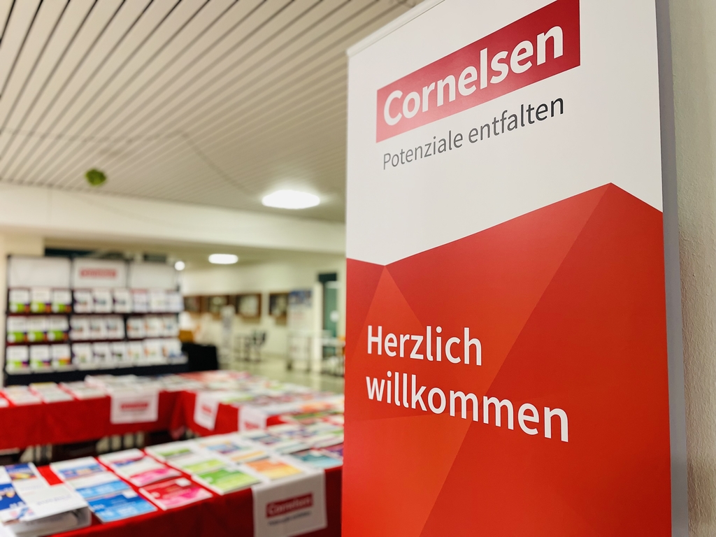 Das Bild zeigt einen Aufsteller von Cornelsen mit der Aufschrift "Herzlich Willkommen". Im Hintergrund sind Regale mit B&uuml;chern und anderen Materialien f&uuml;r den Unterricht zu sehen.