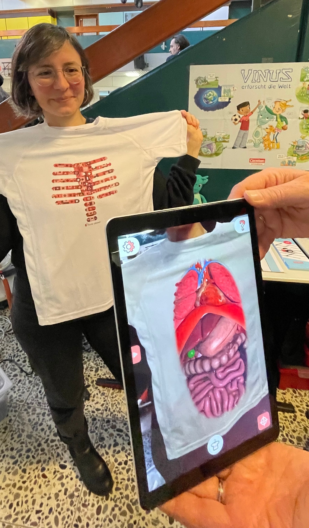 Auf dem Bild h&auml;lt eine Frau das Magic T-Shirt mit einem anatomischen Print, der menschliche Rippen zeigt, vor ihren Oberk&ouml;rper. Daneben wird auf einem Tablet mithilfe der Magic T-Shirt-App eine erweiterte Realit&auml;t (AR)-Darstellung des Inneren eines menschlichen K&ouml;rpers gezeigt.