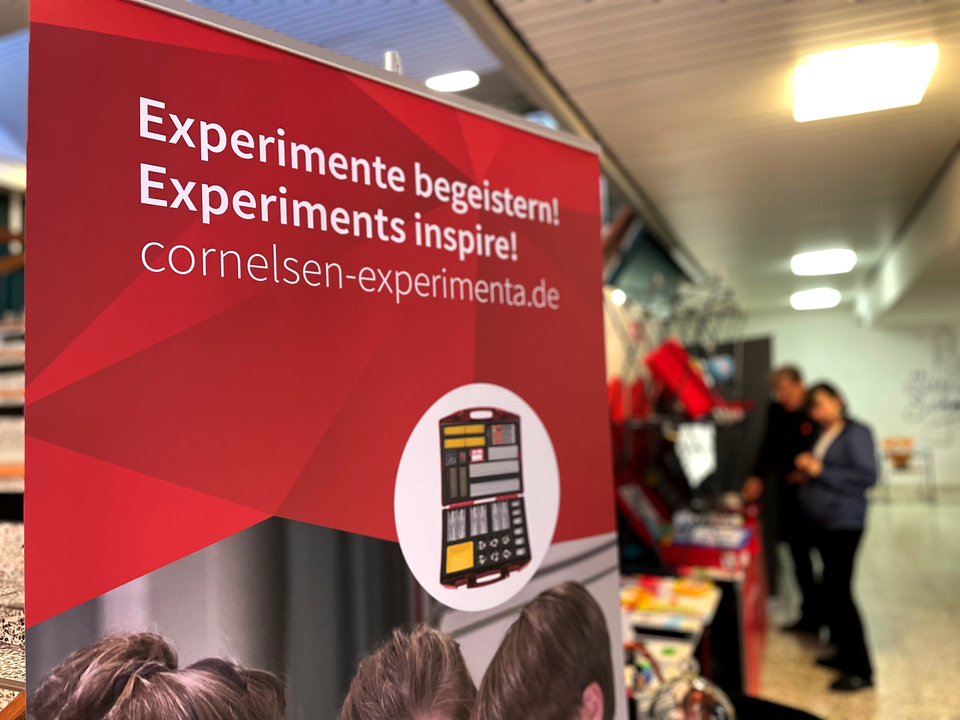 Das Bild zeigt einen Banner von Cornelsen Experimenta auf der MNU Bremerhaven. Es handelt sich um einen Stand f&uuml;r Experimentierbedarf mit der  Botschaft &bdquo;Experimente begeistern!&ldquo; Im Hintergrund stehen zwei Personen und betrachten das Sortiment.
