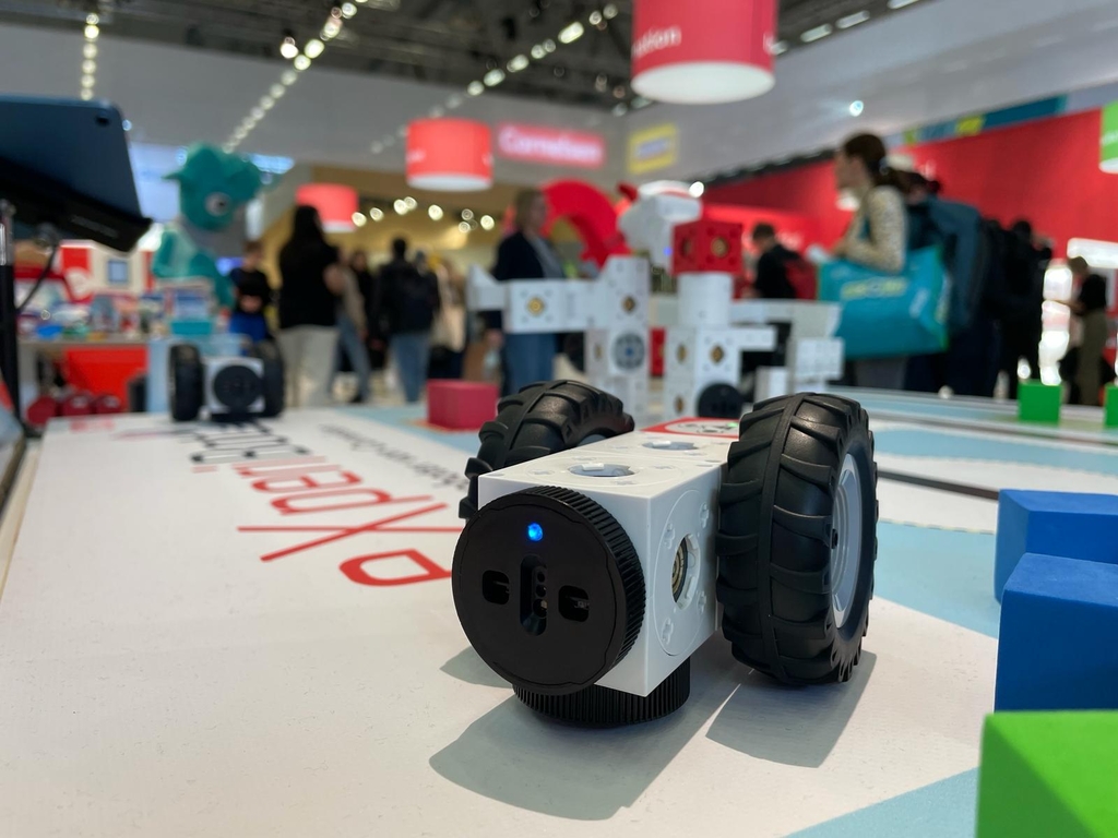 Das Bild zeigt eine Nahaufnahme des eXperiBot Explorers, einem kleinen, wei&szlig;en Roboter mit gro&szlig;en Gummireifen, der auf einer Farbfl&auml;che in einem modernen Setting positioniert ist. Im Hintergrund sind Besucher der didacta 2024 und weitere St&auml;nde zu sehen.