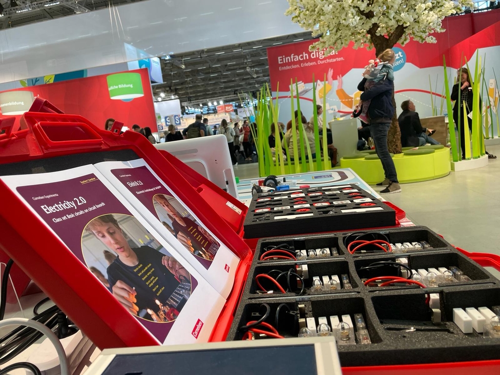 Das Bild zeigt eine rote Kunststoffbox mit Lehrmaterialien zum Thema Elektrizit&auml;t, darunter zwei B&uuml;cher und verschiedene elektronische Bauteile wie Kabel und L&auml;mpchen. Im Hintergrund sind Besucher einer Bildungsveranstaltung zu sehen.