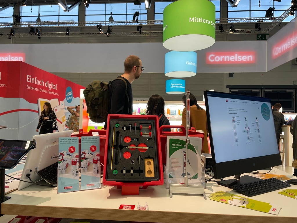 Das Bild zeigt einen Messestand von Cornelsen Experimenta mit Produkten f&uuml;r die Sekundarstufe, darunter der Rote Koffer "Mechanik 2.0" im Vordergrund sowie einem Computer, der ein Experiment zeigt. Im Hintergrund sind farbige Leuchtschilder.