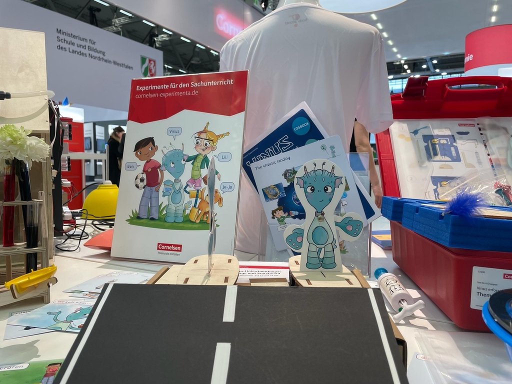 Das Bild zeigt einen Stand mit Experimentiermaterialien f&uuml;r den Sachunterricht, einschlie&szlig;lich einer Brosch&uuml;re mit kindlichen Charakteren und einem roten Kasten mit verschiedenen Experimentier-Utensilien. Au&szlig;erdem sind Illustrationen und Logb&uuml;cher sichtba