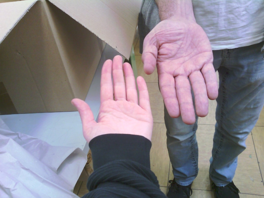 Das Bild zeigt zwei H&auml;nde, die sich gegen&uuml;berstehen, w&auml;hrend im Hintergrund ein Karton sichtbar ist. Die linke Hand geh&ouml;rt zu einer Person mit einem schwarzen Pullover, w&auml;hrend die rechte Hand von einer anderen Person stammt, die ein graues T-Shirt tr&auml;gt.