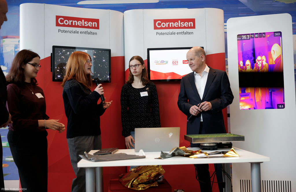 Girls_Day_2024_Bundeskanzler_Wärmebildkamera / Drei junge Schülerinnen und Bundeskanzler Olaf Scholz stehen vor einem Messestand von Cornelsen mit dem Slogan "Potenziale entfalten". Eine der Schülerinnen hält ein Mikrofon und spricht, während die anderen 