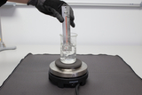 Das Bild zeigt ein Becherglas mit einer klaren Flüssigkeit, das auf einem Heizgerät steht. Eine Person in Handschuhen hält ein Thermometer in das Glas, um die Temperatur zu messen.