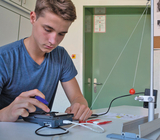 Das Bild zeigt einen Jugendlichen, der an einem Experimentiergerät arbeitet, wobei er einen Taschenrechner und einige elektrische Verbindungen benutzt. Im Hintergrund ist ein Pendel mit einer roten Kugel zu sehen, was auf eine physikalische Messung hindeu