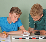 Das Bild zeigt zwei Jungen, die sich an einem Experiment mit einem optischen Gerät beteiligen. Sie arbeiten an einem Tisch, auf dem eine experimentelle Apparatur sowie Arbeitsblätter liegen, während sie die Ergebnisse genau beobachten und aufzeichnen.