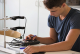 Das Bild zeigt einen Schüler, der an einem Tisch mit verschiedenen Experimentiermaterialien arbeitet. Vor ihm befindet sich ein Gerät, das mit Kabeln verbunden ist, während er Notizen macht und einen Stift in der Hand hält.