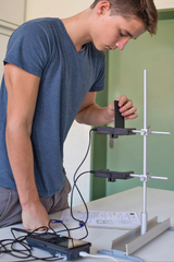 Das Bild zeigt einen Schüler, der an einem Experimentiergerät arbeitet, während er Sensoren hält. Auf dem Tisch liegen technische Geräte sowie ein Arbeitsblatt, das wahrscheinlich zur Durchführung des Experiments dient.