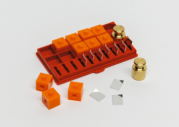 Das Bild zeigt ein Experimentierset mit orangefarbenen Blöcken, zwei goldenen Gewichten und einzelnen metallischen Plättchen auf einer roten Ablage. Diese Materialien sind für pädagogische Zwecke im deutschen Bildungswesen konzipiert, um physikalische Exp