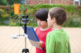 Auf dem Bild sind zwei Kinder, ein Junge und ein Mädchen, die an einem Experimentierstand arbeiten. Sie scheinen eine Wetterstation zu beobachten, während das Mädchen einen Clip-Board mit Notizen in der Hand hält.