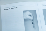 Das Bild zeigt eine Hand, die einen Magneten hält, während er an zwei Metallgewichten befestigt ist. Der Text daneben betont, dass "Magnete Kraft haben", und vermittelt so das Konzept der magnetischen Anziehung.