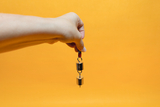 Das Bild zeigt eine Hand, die ein Experimentiergerät mit zwei metallischen Gewichten hält, die an einem roten Kabel befestigt sind. Der Hintergrund ist orange, was den Fokus auf das Experimentiermaterial verstärkt und es für den Bildungsbereich attraktiv 