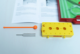Das Bild zeigt ein Experimentier-Kit mit einem gelben Schaltblock, einem Stimmgabel, einem roten Stab und einer orangefarbenen Kugel. Daneben liegt ein Anleitungshandbuch, das die Materialien und Vorgehensweisen für verschiedene Versuche im Bildungsbereic