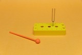 Das Bild zeigt ein gelbes Experimentiergerät mit mehreren Löchern, in das eine Stimmgabel eingesetzt ist. Neben dem Gerät liegt ein orangefarbener Kunststoffstab, wahrscheinlich für die Durchführung von Experimenten im Bildungsbereich.
