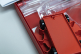Das Bild zeigt eine rote Experimentierbox, die einen Fahrzeugbausatz enthält. Ein rotes Fahrzeugteil mit schwarzen Rädern ist sichtbar, während sich transparente Kunststoffteile und eine Anleitung im Hintergrund befinden.