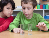 Das Bild zeigt zwei Kinder, die aufmerksam vor zwei Messbechern mit Wasser sitzen. Der Junge in grün deutet auf einen der Becher, während das Mädchen in rot gespannt zuschaut, was auf ein Experiment oder eine Beobachtung hinweist.