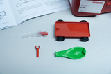 Das Bild zeigt verschiedene Experimentiermaterialien, darunter ein rotes Fahrzeug mit Rädern, eine grüne Ballon und weitere Komponenten, die für experimentelle Zwecke genutzt werden können. Im Hintergrund liegt eine Anleitung, die die Verwendung dieser Te