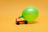 Das Bild zeigt ein kleines Fahrzeug, auf dem ein grüner Luftballon montiert ist. Das Fahrzeug steht auf einer orangefarbenen Fläche und demonstriert wahrscheinlich ein Prinzip der Luftdruck- oder Bewegungslehre.