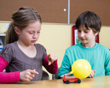 Das Bild zeigt zwei Kinder, die an einem Experiment mit einem kleinen, rot lackierten Fahrzeug arbeiten, das mit einem gelben Ballon ausgestattet ist. Die Kinder scheinen interessiert und engagiert in das Experiment involviert zu sein, während sie die Mec