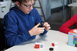 Das Bild zeigt einen Schüler, der ein schwarzes Element in der Hand hält und es mit einem Konstruktionsspielzeug auf dem Tisch kombiniert. Mehrere bunte Module liegen auf dem Tisch, während ein weiterer Schüler im roten Shirt beobachtet.