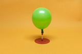 Das Bild zeigt eine grüne Luftballon-Dekoration, die auf einem stabilen, runden roten Standfuß montiert ist. Der Hintergrund ist ein sanftes Gelb, was die lebendige Farbe des Ballons hervorhebt und eine freundliche Atmosphäre schafft.