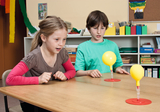 Das Bild zeigt zwei Kinder, die gespannt auf zwei gelbe Luftballons schauen, die auf einem Tisch stehen. Die Ballons sind durch transparente Schläuche verbunden, und die Kinder scheinen an einem Experiment teilzunehmen, das wissenschaftliche Konzepte verm