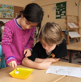 Das Bild zeigt zwei Kinder, die gemeinsam an einem Experiment arbeiten. Das Mädchen in einem lila Pullover taucht einen Gegenstand in eine gelbe Schale, während der Junge aufmerksam neben ihr sitzt und ein Arbeitsblatt betrachtet.