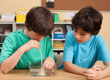 Das Bild zeigt zwei Jungen, die in einem Experimentier-Set arbeiten. Einer der Jungen verwendet ein Röhrchen, um eine kleine gelbe Kugel in einem Wasserbehälter zu bewegen, während der andere aufmerksam zuschaut.