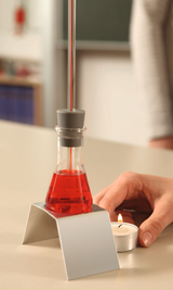 Auf dem Bild ist ein Reagenzglas mit roter Flüssigkeit zu sehen, das auf einem stabilen Ständer steht. Neben dem Glas befindet sich eine brennende Teekerze, während eine Hand das Glas hält, was einen Experimentieraufbau im Bildungsbereich zeigt.