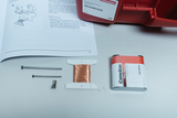 Auf dem Bild sind verschiedene Experimentiermaterialien zu sehen, darunter eine Spule mit Kupferdraht, zwei Nägel, ein Anschlussstück und eine Batterie. Außerdem liegt ein Lehrbuch mit Anweisungen zu Stromkreisen daneben.