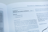 Das Bild zeigt eine Anleitung zu Experimenten mit einer Lampe und einer Batterie aus einem Lehrbuch für das deutsche Bildungswesen. Es listet das benötigte Material auf, einschließlich einer Flachbatterie und einer Glühlampe, sowie wichtige Hinweise zur D
