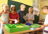 Das Bild zeigt mehrere Kinder, die an einem Experimentiertisch stehen und mit verschiedenen Materialien aus einer grünen Schaumstoffbox arbeiten. Sie scheinen konzentriert zu sein, während sie sich mit ihren Projekten im Klassenzimmer befassen.