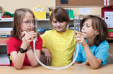 Das Bild zeigt drei Kinder, die gespannt zwei mit Wasser gefüllte Glaskolben halten, die durch einen Schlauch miteinander verbunden sind. Sie experimentieren und beobachten wahrscheinlich ein physikalisches Phänomen, während eine freundliche Lernatmosphär
