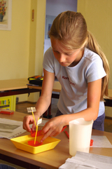 Das Bild zeigt ein Mädchen, das konzentriert an einem Experiment arbeitet, indem es eine gelbe Schale mit roter Flüssigkeit und mehreren beschrifteten Stäbchen nutzt. Im Hintergrund sind Tische und eine Unterrichtsatmosphäre zu erkennen, die für schulisch