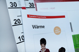 Das Bild zeigt eine Lehrmaterialien mit dem Titel "Wärme" von Cornelsen Experimenta, die sich auf Experimente zum Thema Wärme konzentriert. Im Hintergrund ist ein Thermometer sichtbar, das verschiedene Temperaturgrade anzeigt.