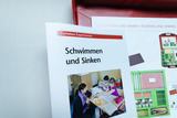 Das Bild zeigt eine Broschüre mit dem Titel „Schwimmen und Sinken“ aus einem Experimentierset für den Bildungsbereich. Darin sind Kinder zu sehen, die zusammen mit einer Lehrperson an einem Tisch experimentieren und lernen.