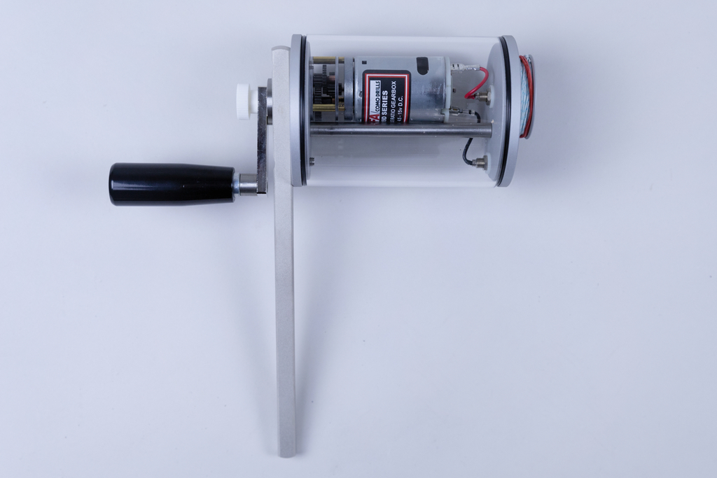 Das Bild zeigt ein transparentes Gerät mit einem Handgriff und einem Motor im Inneren. Es scheint für Experimente im Bildungsbereich konzipiert zu sein, möglicherweise zur Demonstration von mechanischen Bewegungen oder Energieumwandlungen.