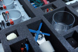 Das Bild zeigt eine kompakte Aufbewahrung mit verschiedenen Experimentiermaterialien, darunter ein kleiner blauer Propeller, elektrische Bauteile und ein Glasbehälter. Diese Materialien sind ordentlich in Fächer angeordnet, um einen strukturierten Zugang 